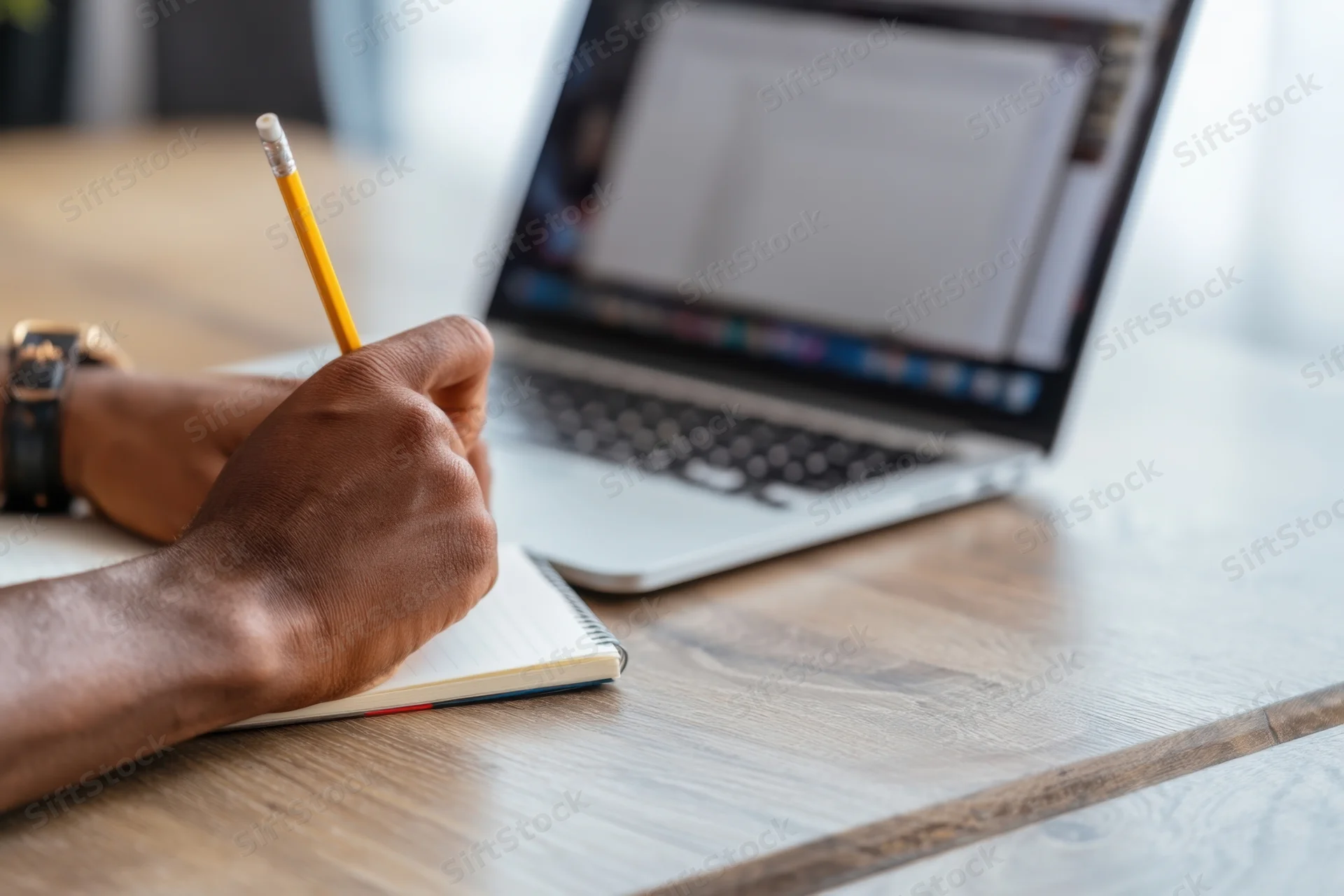 Man writing in notebook with pencil beside open laptop on wooden desk
