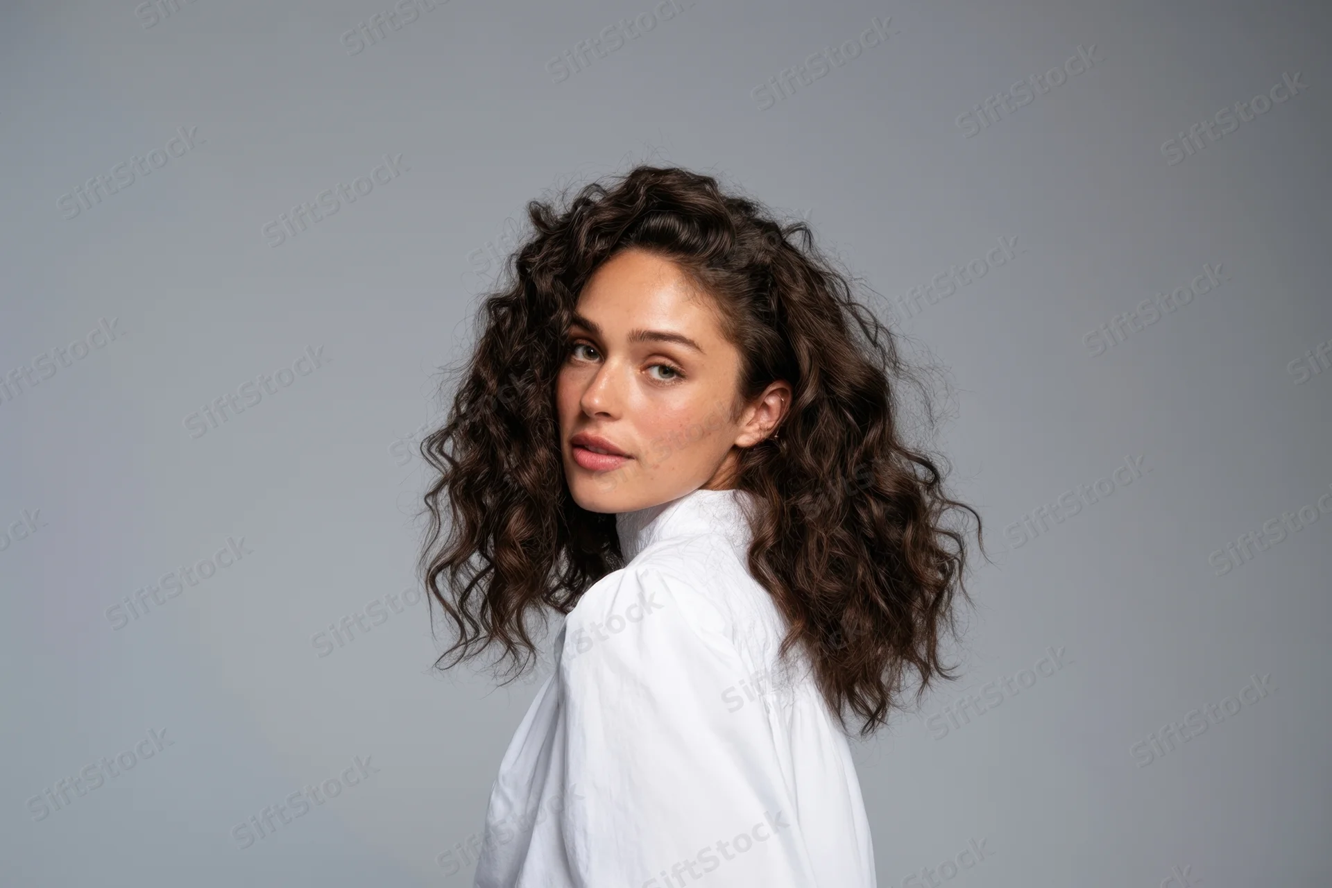 Young woman with curly hair in white shirt turning to look at camera in studio setting