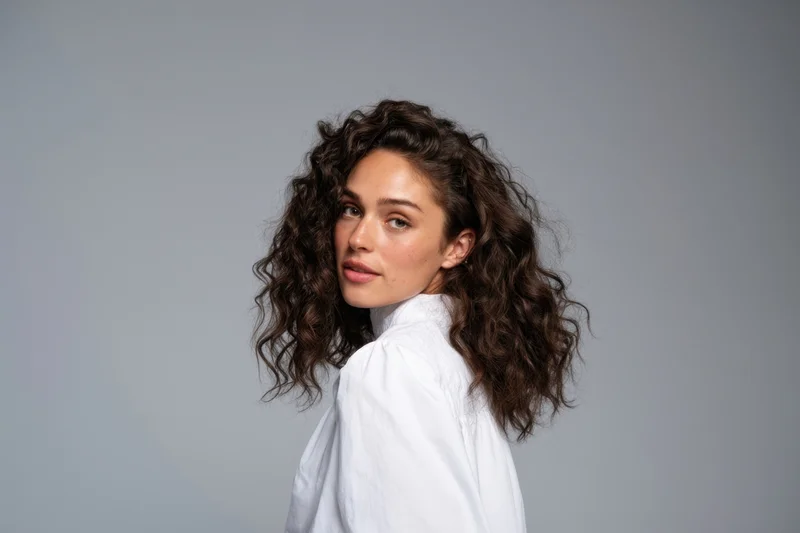 Young woman with curly hair in white shirt turning to look at camera in studio setting