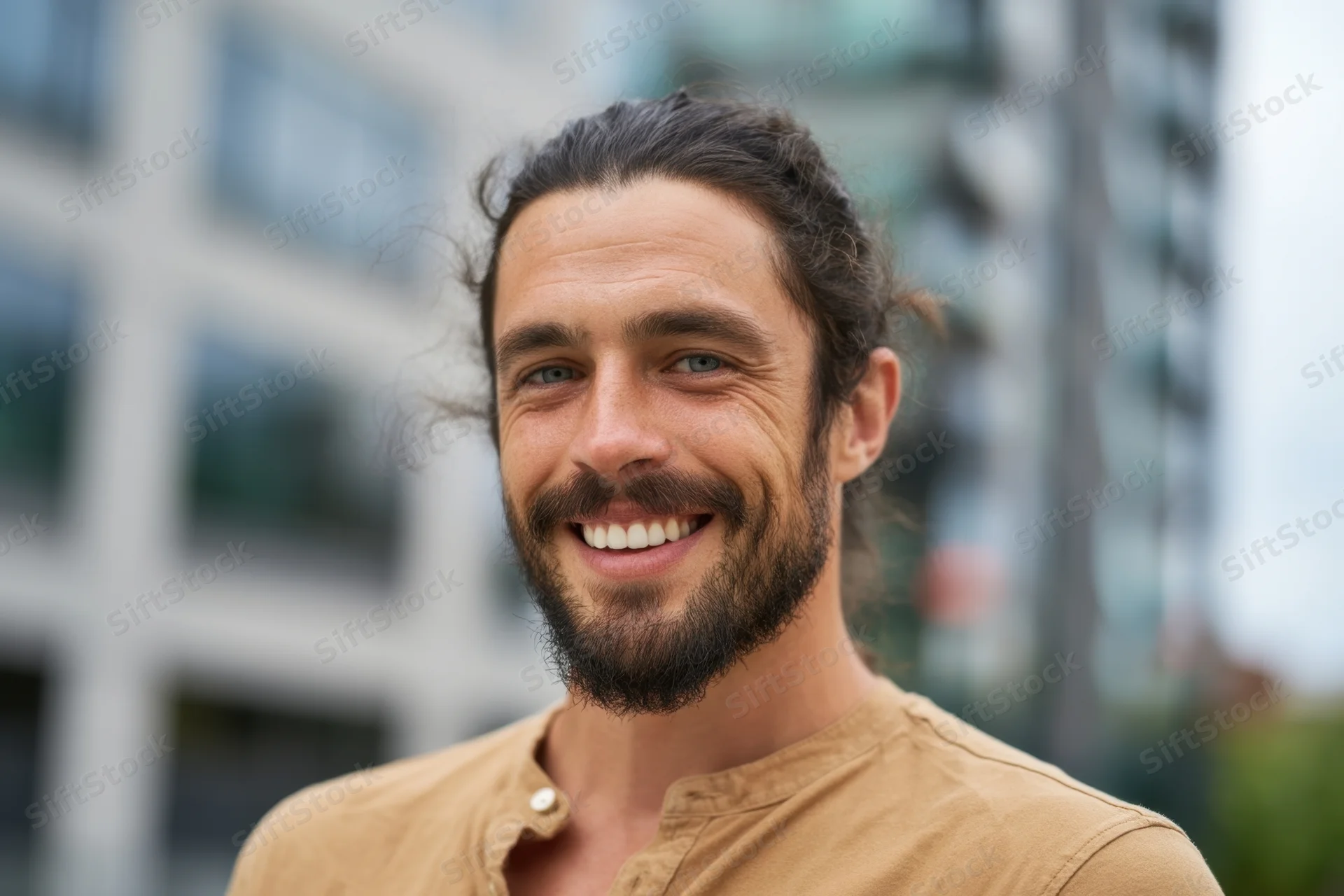 Smiling man with dark hair tied back and full beard outdoors in city setting