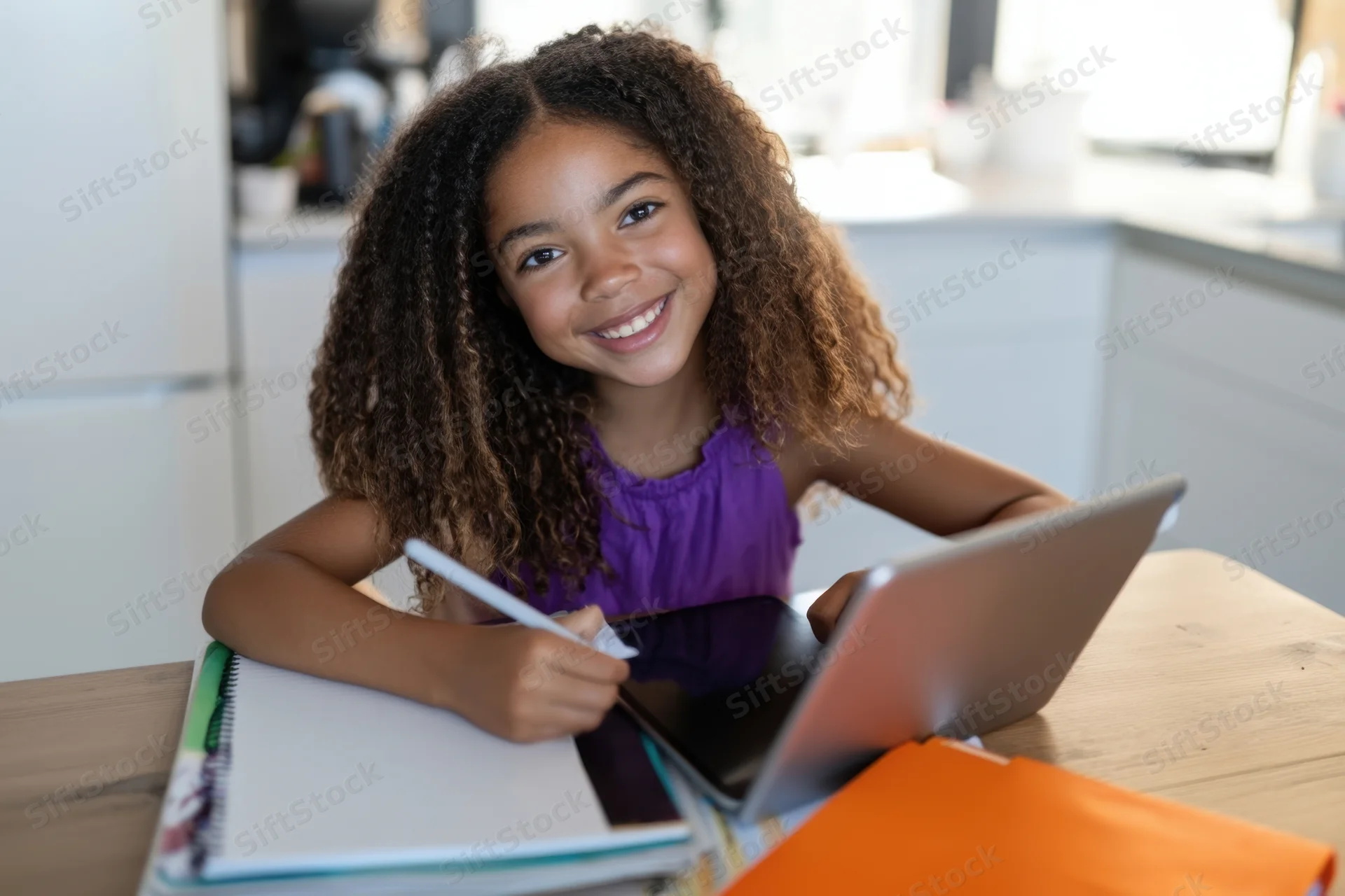 Young girl smiling while writing in a notebook with a tablet on a wooden kitchen table