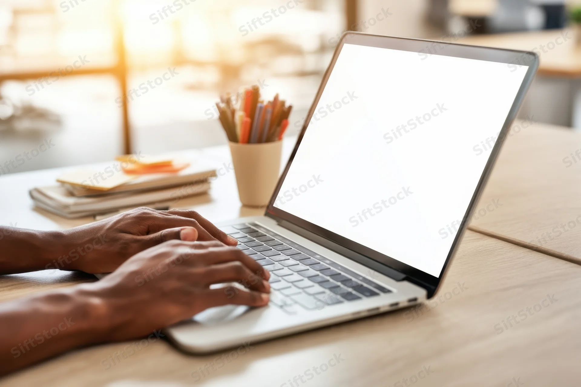 Hands typing on a silver laptop with blank white screen on a wooden desk in a sunlit workspace