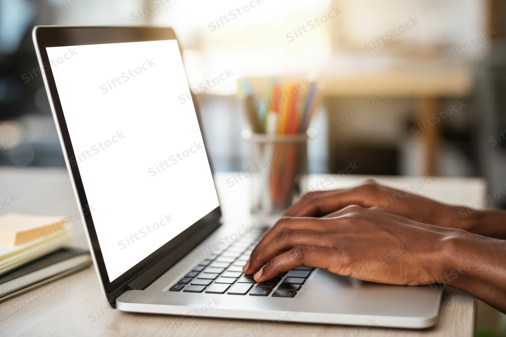 Hands typing on a silver laptop with blank screen on a wooden desk in a sunlit workspace