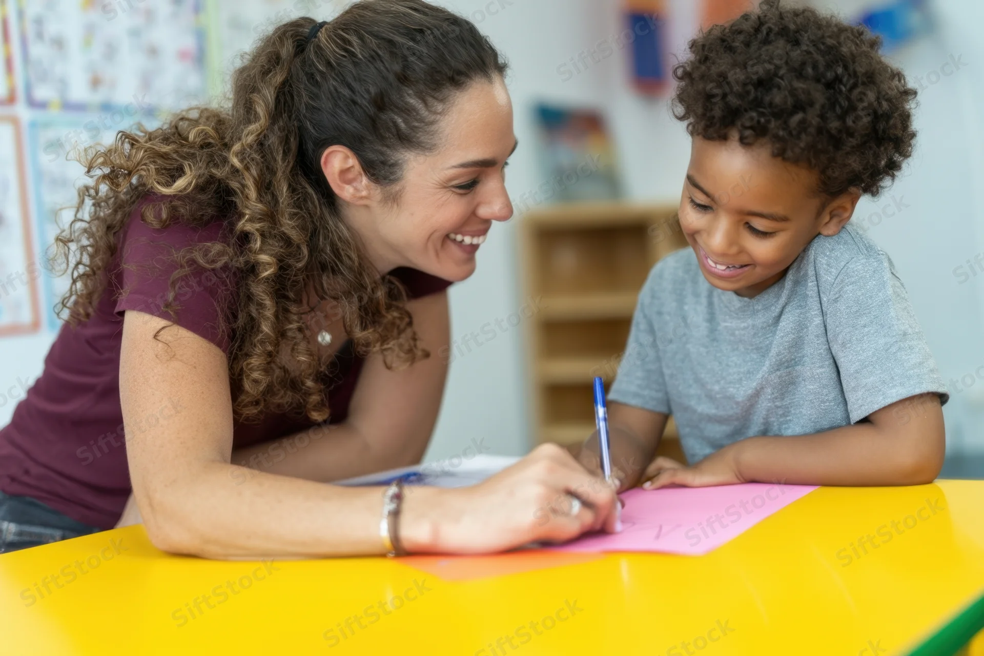 Teacher smiling while writing with a young boy at a yellow desk in a classroom
