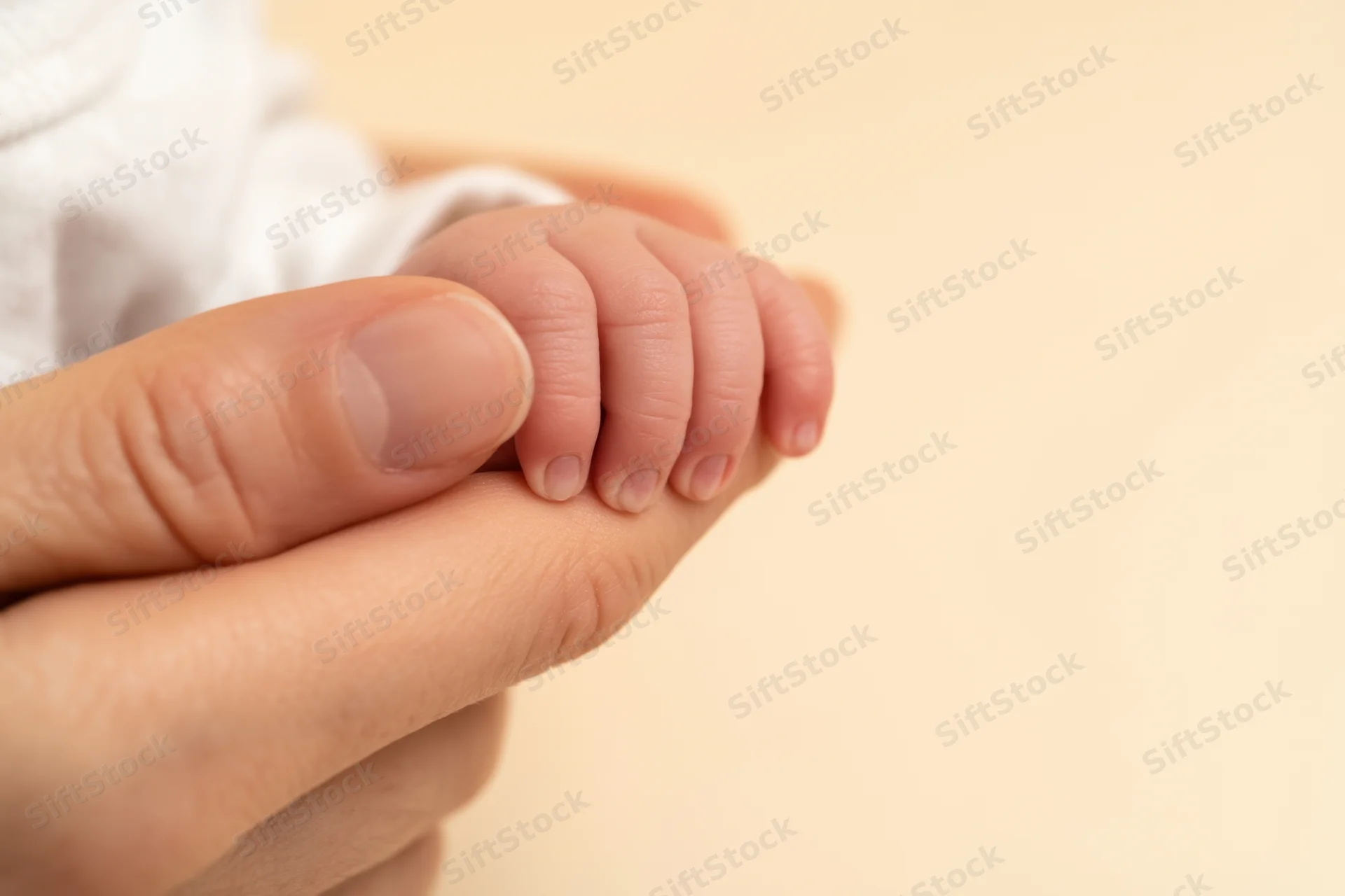 Baby's tiny hand gripping an adult finger against a soft beige background