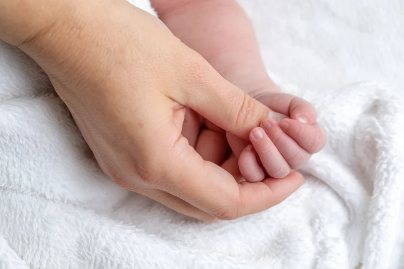 Adult hand gently holding newborn baby's hand on white blanket