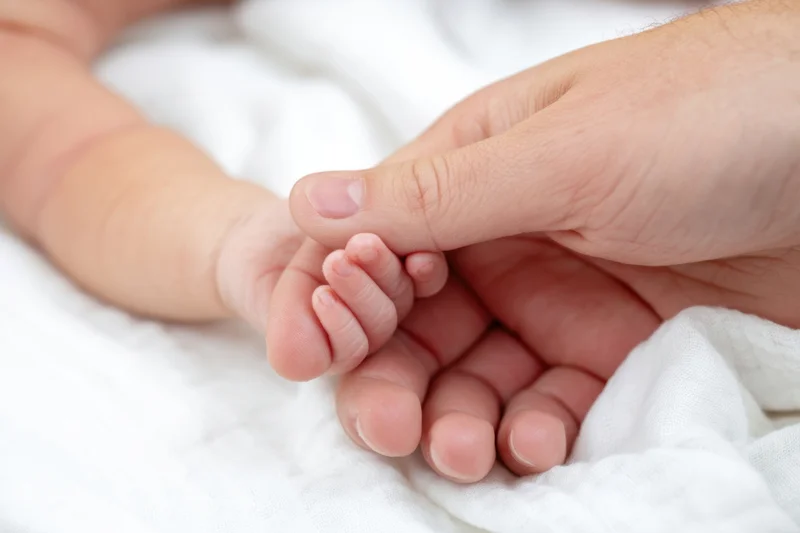 Adult hand gently holding a newborn baby's hand on a white blanket