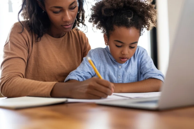 Woman helping young girl with homework at wooden table using laptop and pencil