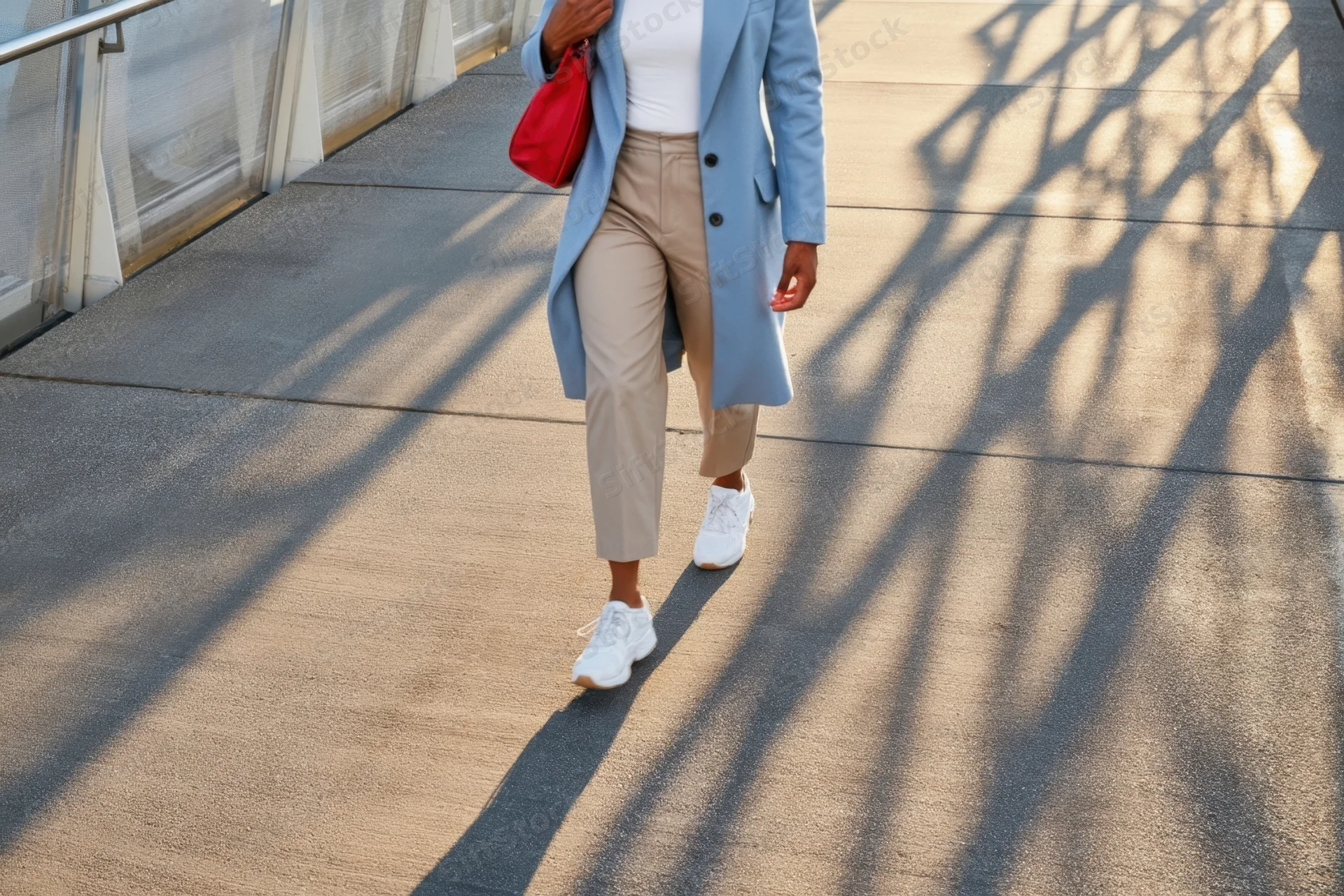 Woman walking on a sunlit concrete walkway in a blue coat and beige trousers