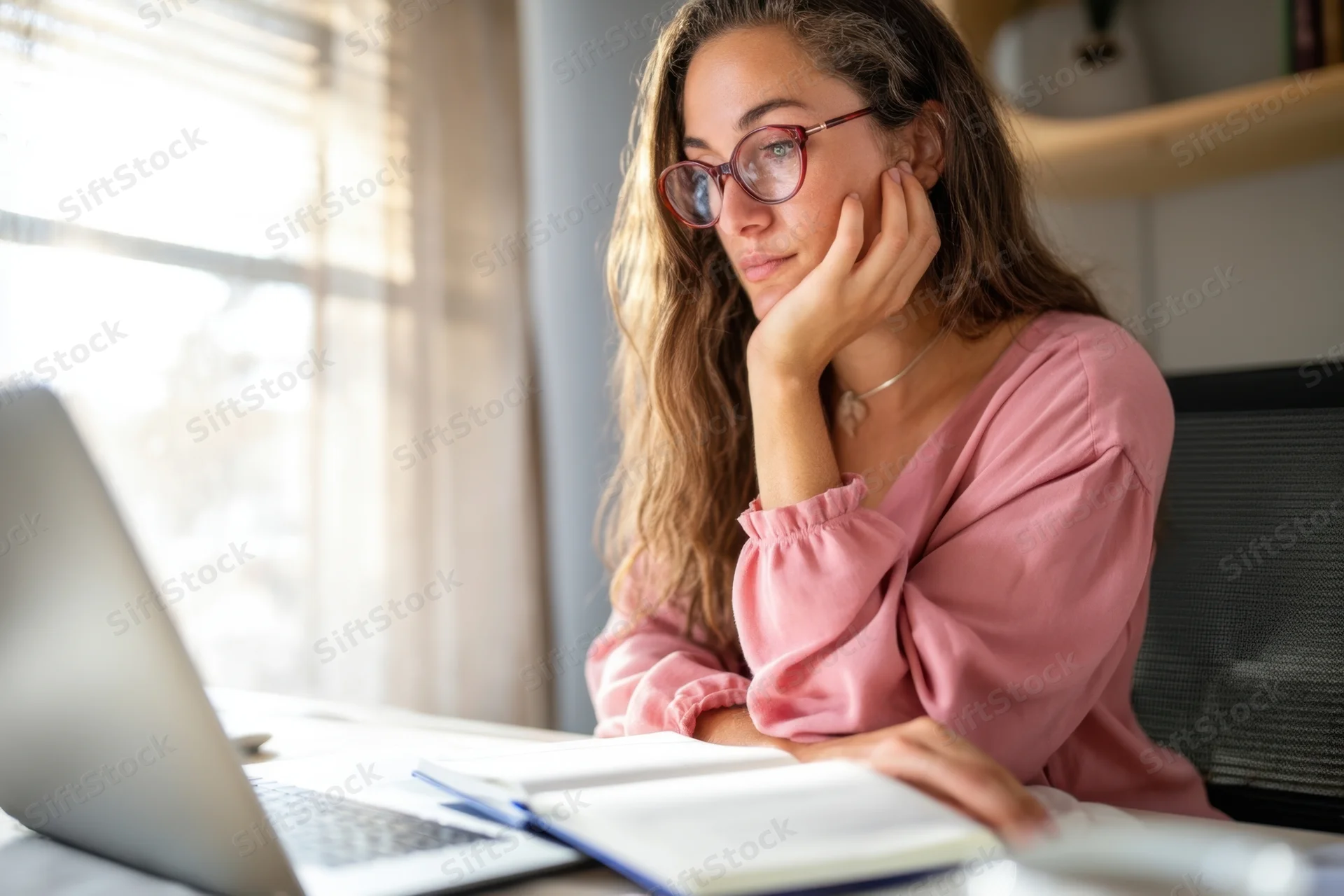 Woman in pink blouse studying at desk with laptop and notebook, hand on chin, focused on screen