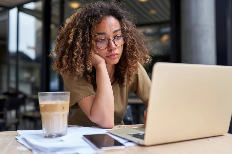 Woman with curly hair and round glasses working on laptop at cafe table with coffee and documents