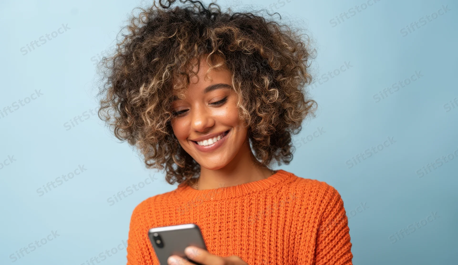 Woman smiling while using smartphone in orange knit sweater against light blue background