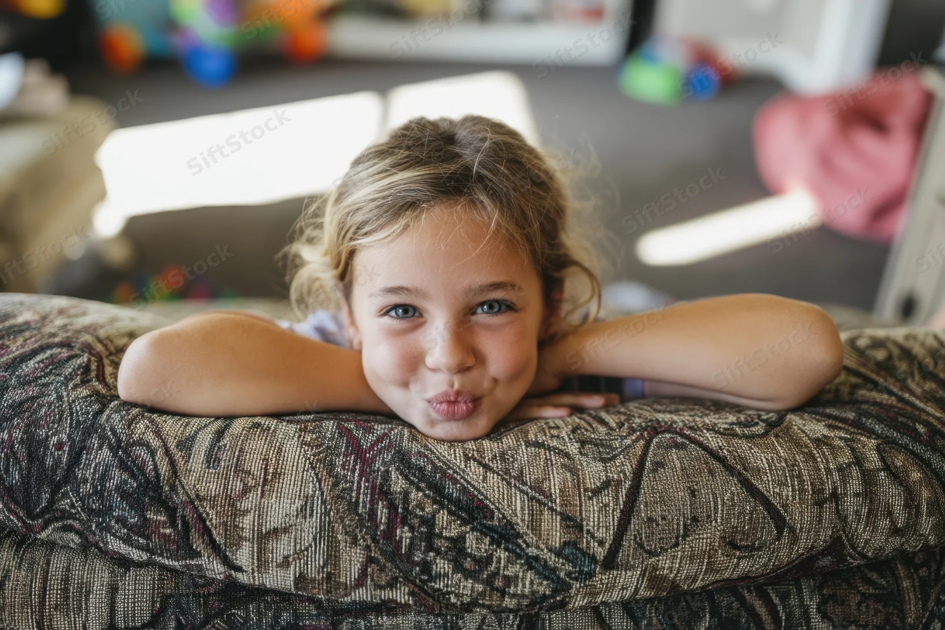 Young girl lying on patterned couch with arms resting on back, making playful pouting expression indoors