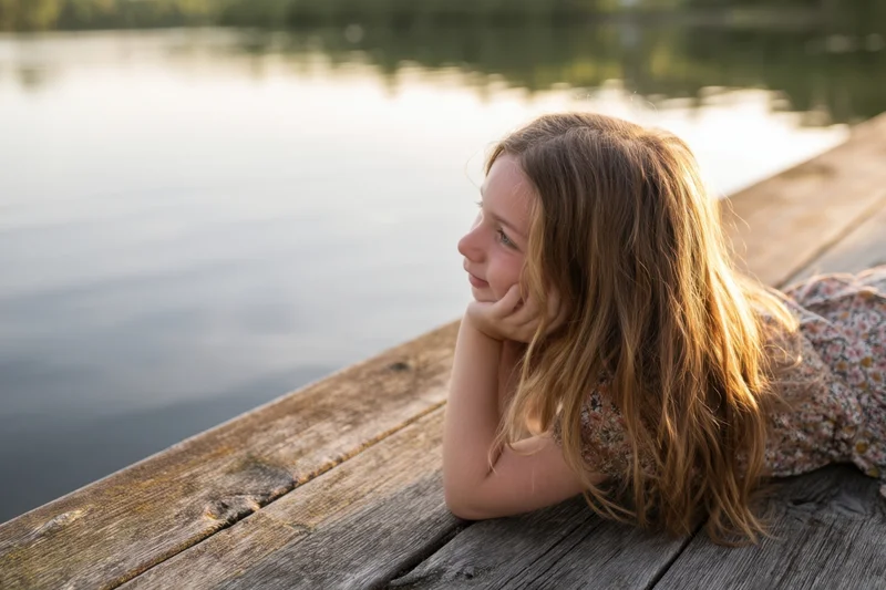 Young girl lying on a wooden dock by a calm lake, gazing into the distance at golden hour