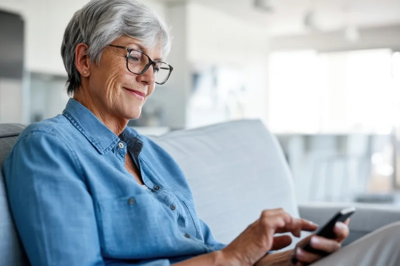 Senior woman smiling while using smartphone on couch in living room