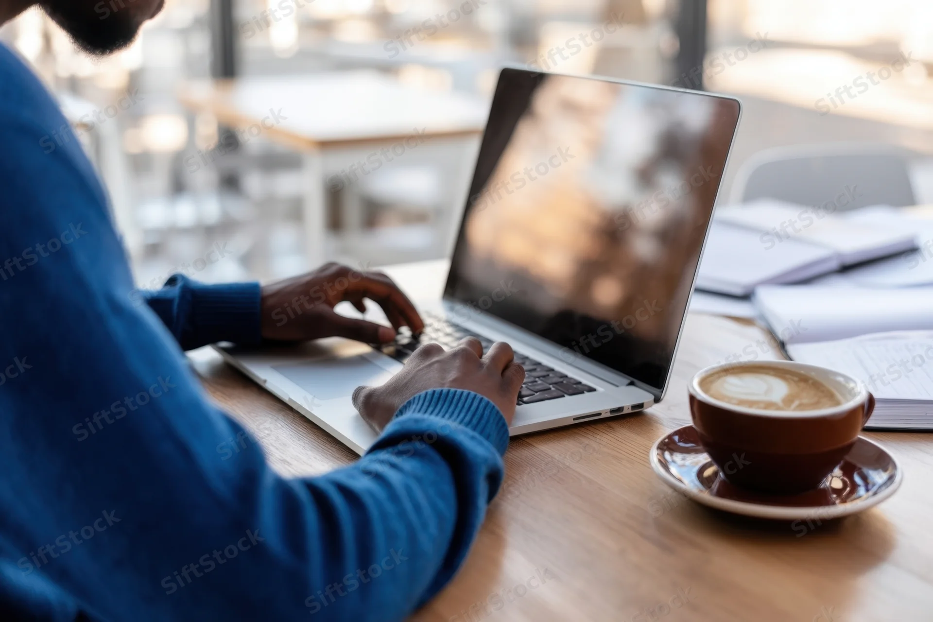 Man typing on laptop at wooden table in cafe with coffee cup and notebook nearby