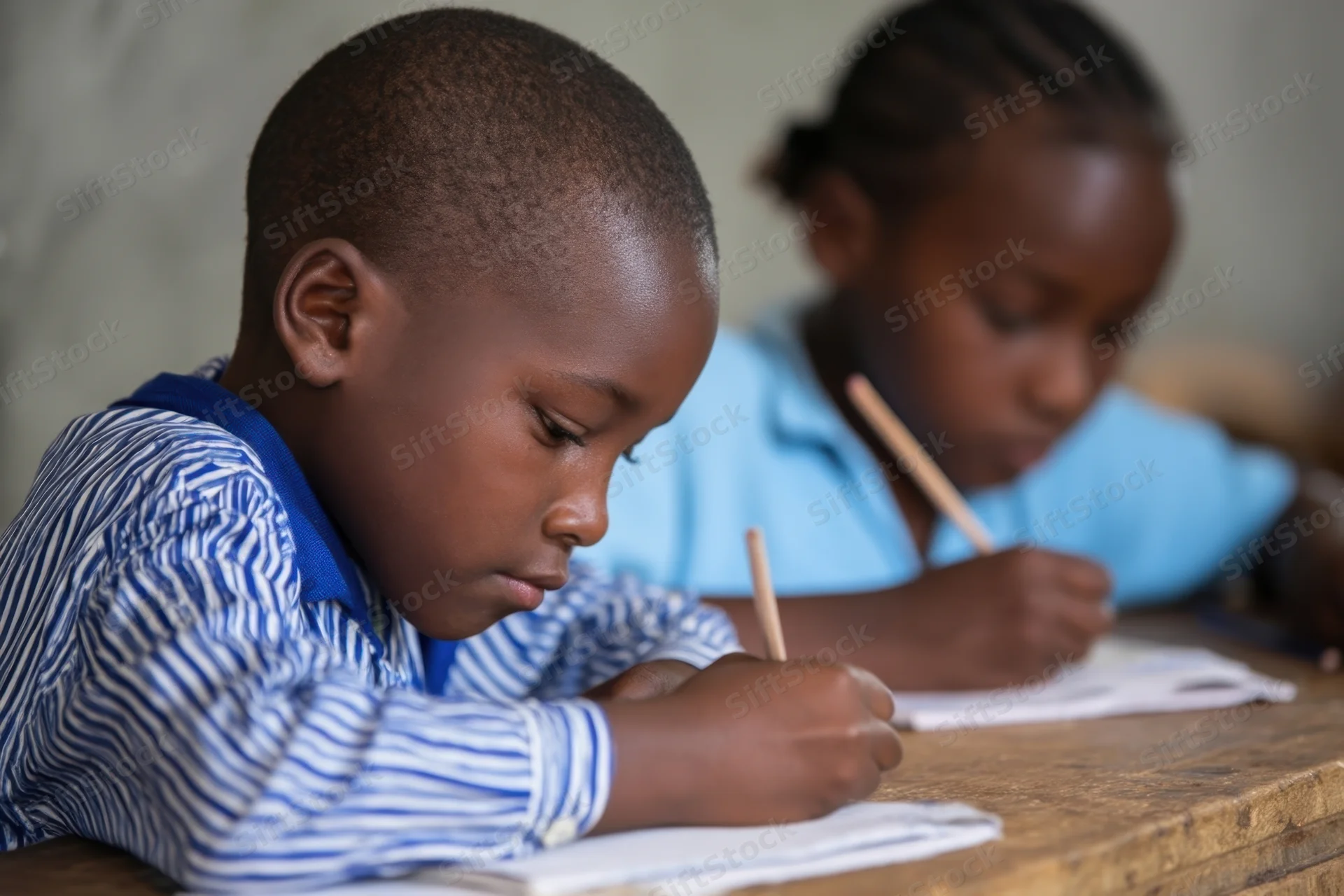 Two children writing in notebooks at a wooden table in a classroom
