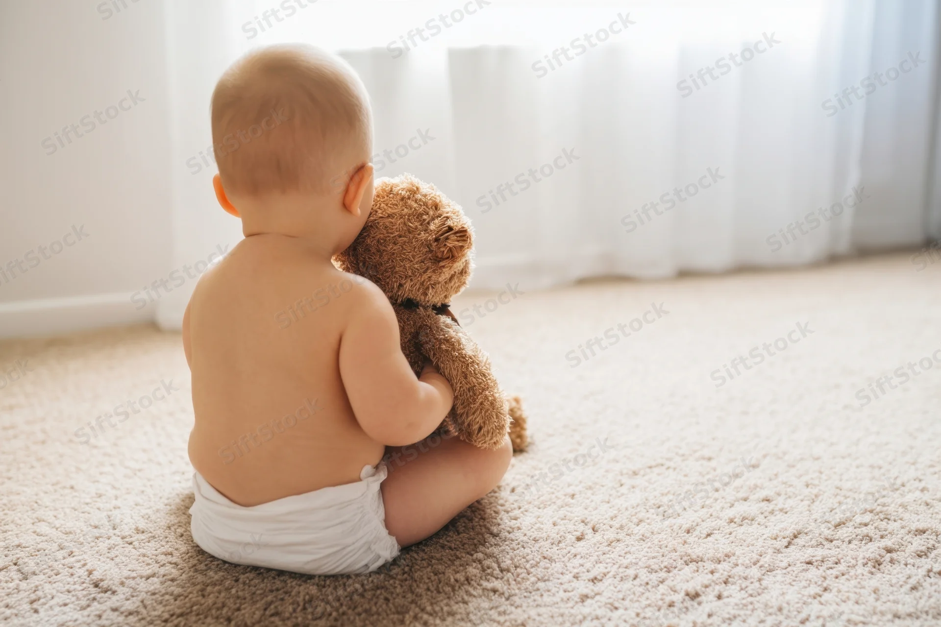 Baby sitting on beige carpet hugging teddy bear facing away from camera