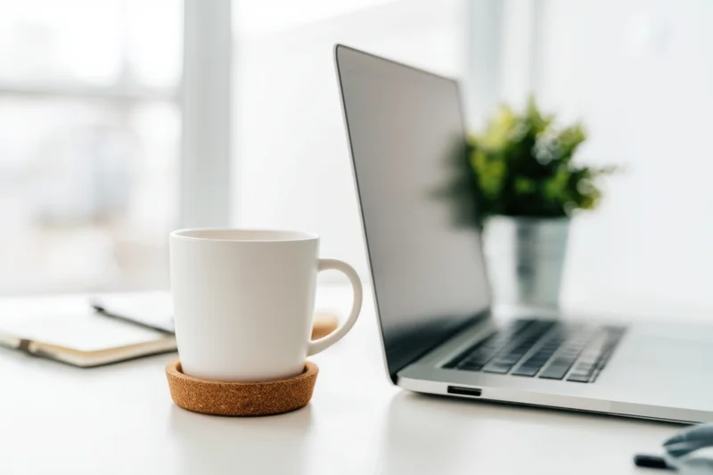 Silver laptop open on white desk beside white mug on cork coaster with notebook and plant in background