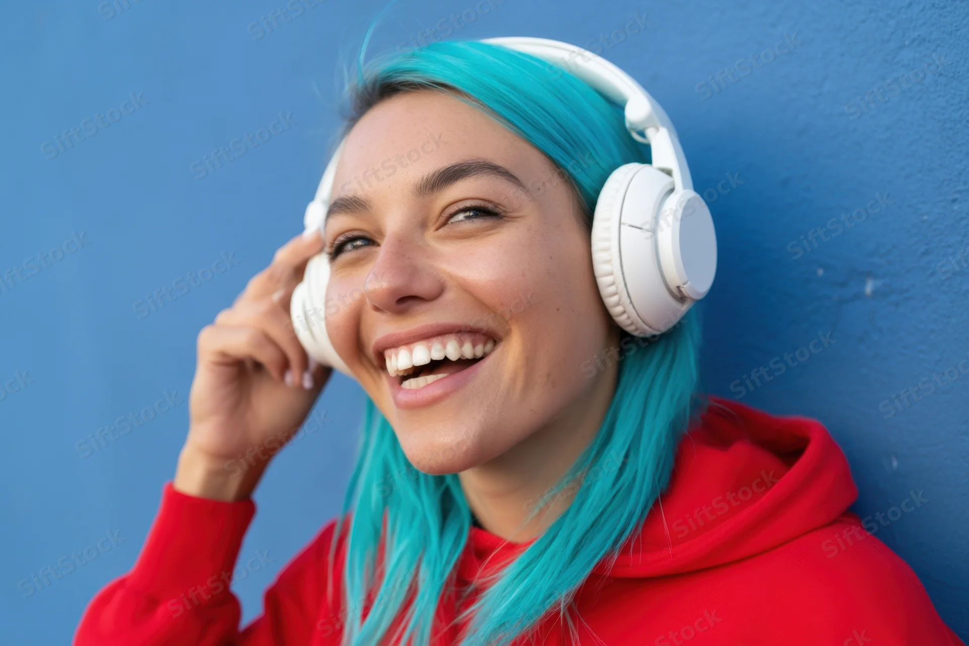 Smiling woman with turquoise hair wearing red hoodie and white headphones against blue wall