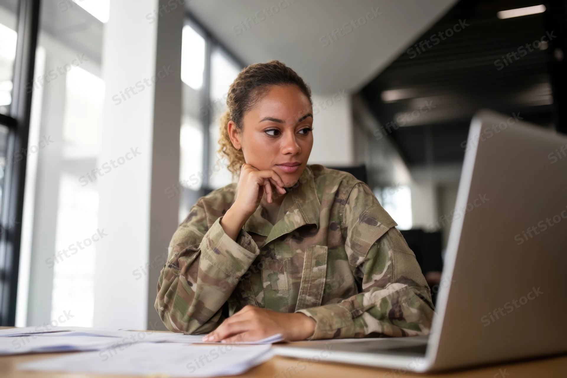 Female soldier in camouflage uniform working at a desk with laptop and documents in an office