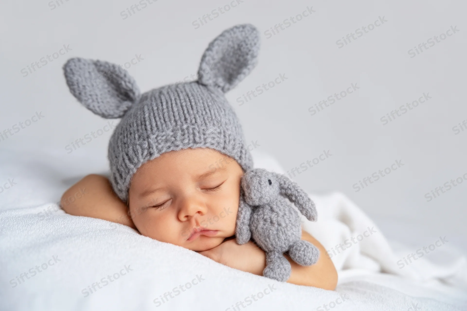 Newborn baby sleeping peacefully in a gray knitted bunny hat with a matching stuffed rabbit toy on a white blanket