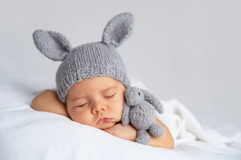 Newborn baby sleeping peacefully in a gray knitted bunny hat with a matching stuffed rabbit toy on a white blanket