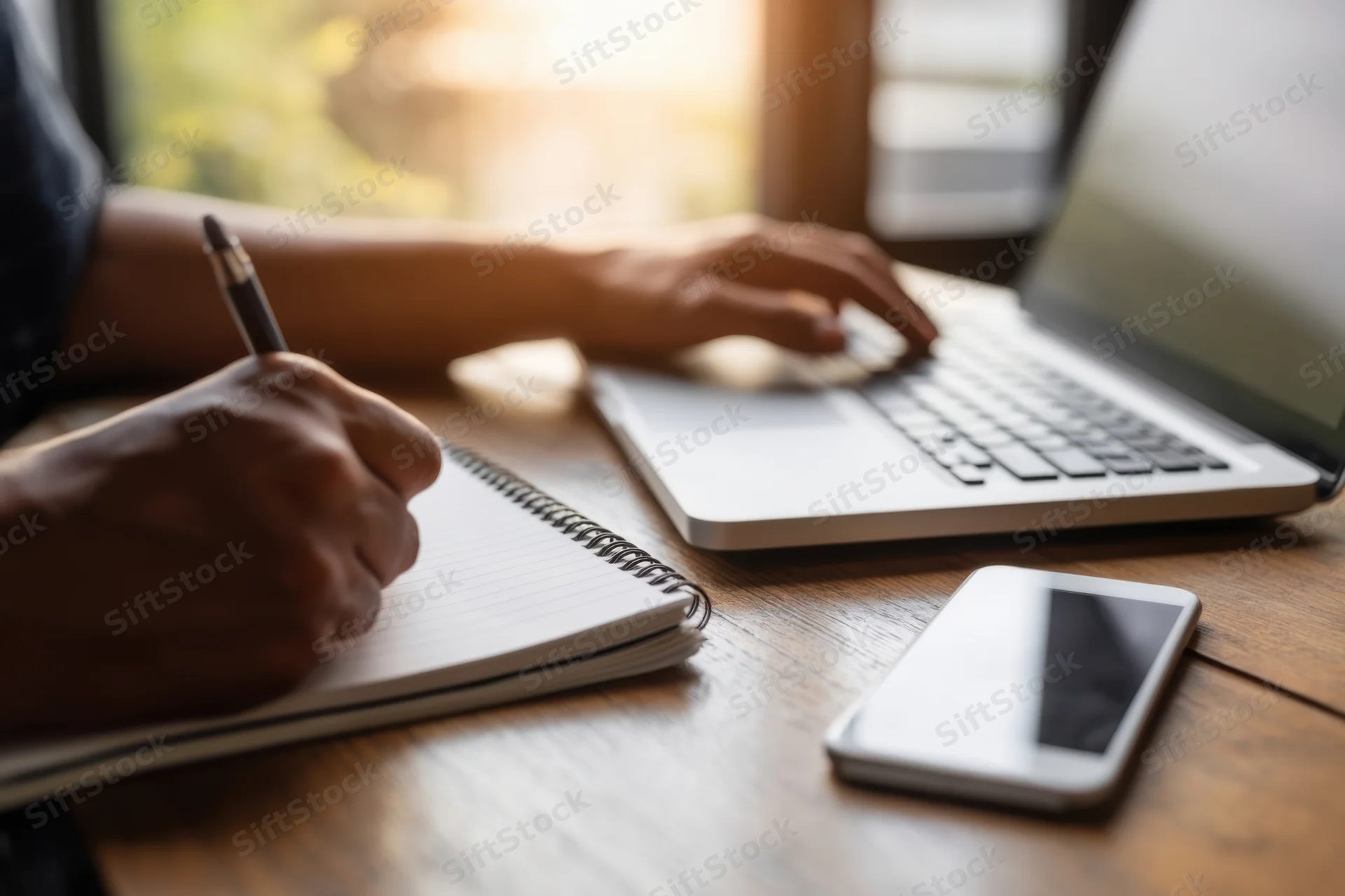 Person writing in spiral notebook beside open laptop on wooden desk
