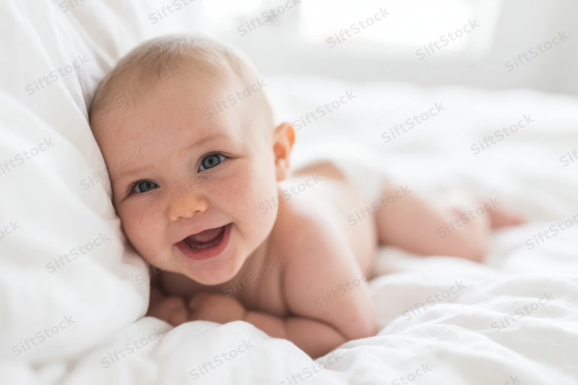 Smiling baby lying on tummy on white bedding in soft natural light