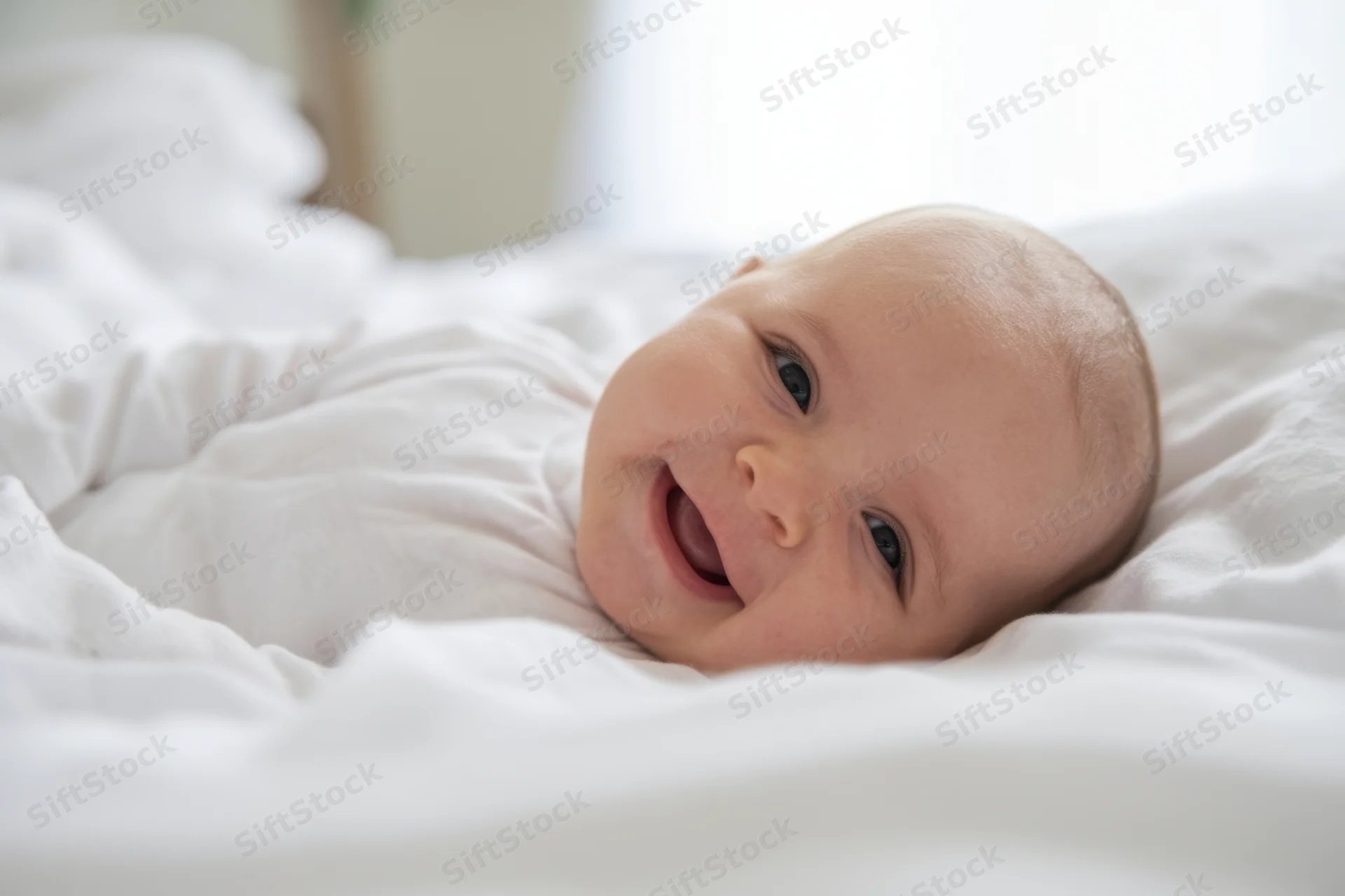Smiling baby lying on white bedding in soft indoor light