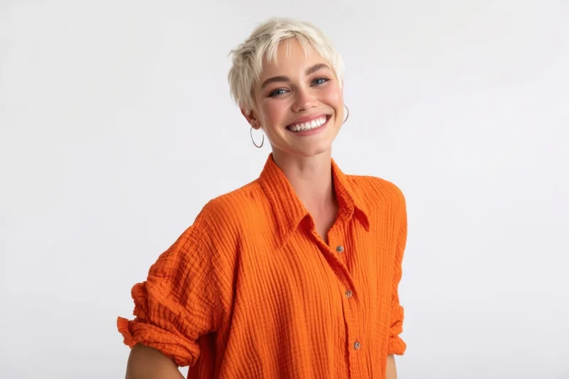 Smiling woman with short platinum-blonde hair wearing an orange ribbed shirt in a studio setting