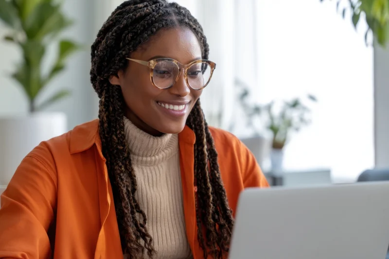 Smiling woman with braided hair and glasses working on laptop at office desk