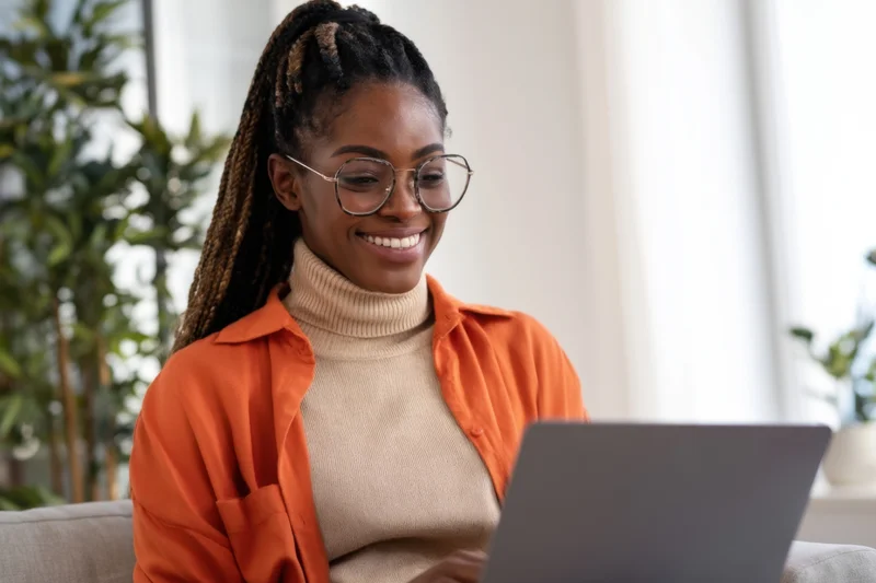 Smiling woman with braided hair and glasses using laptop at home