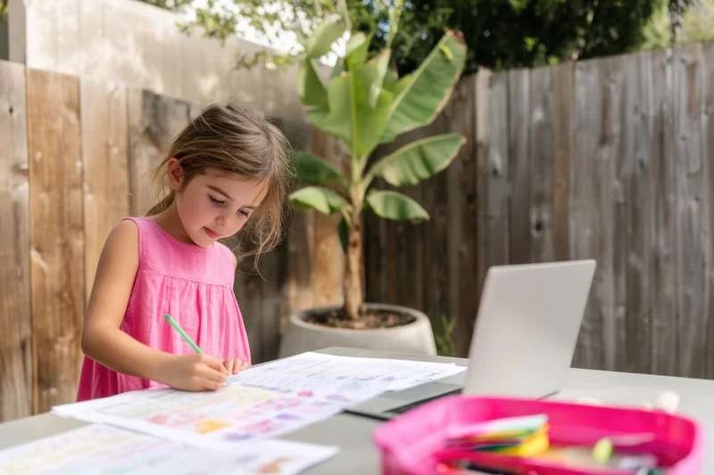 Young girl drawing on paper at outdoor table with laptop and craft supplies nearby