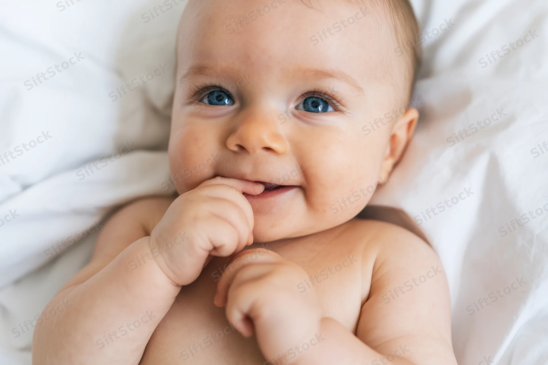 Baby lying on white bedding with hand near mouth, smiling at camera