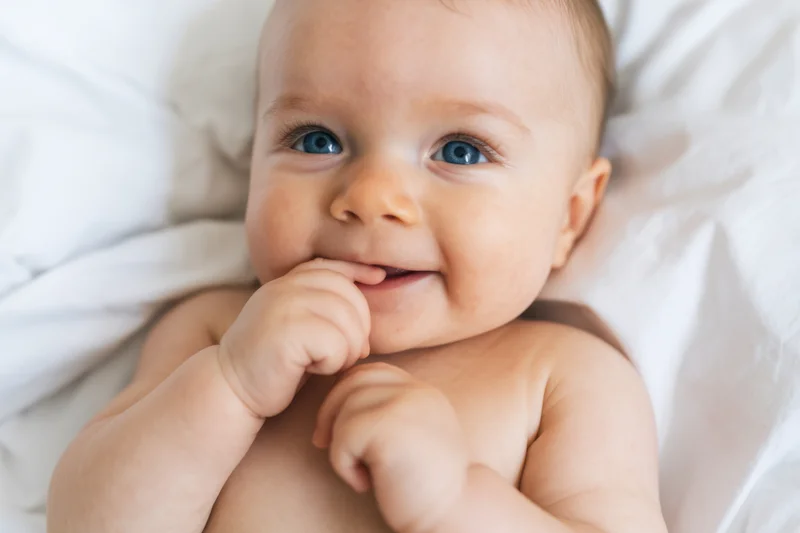 Baby lying on white bedding with hand near mouth, smiling at camera