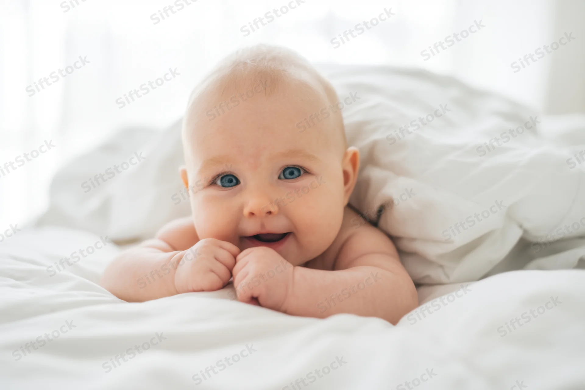 Baby smiling with blue eyes on white bedding in soft natural light
