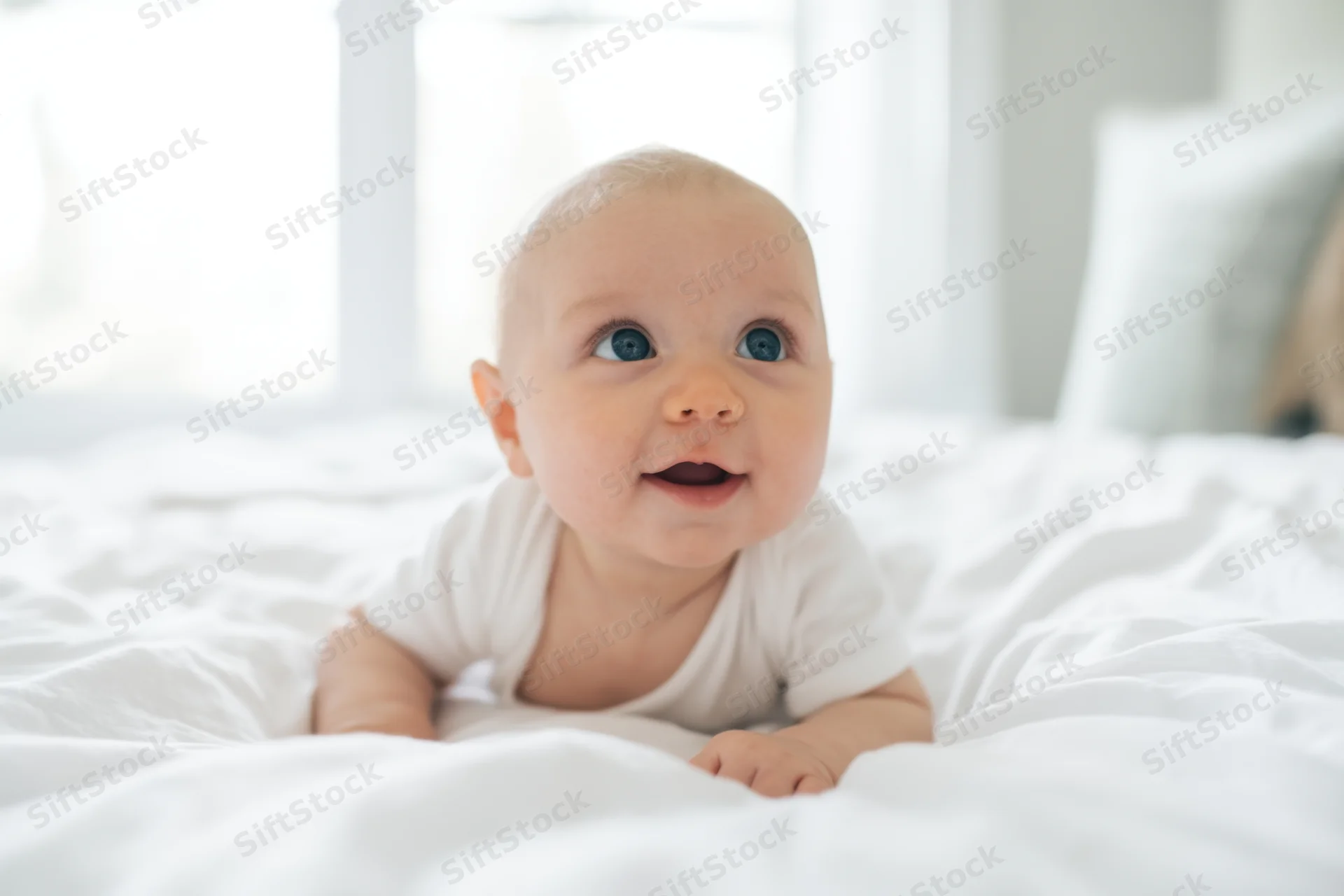 Baby lying on white bed in onesie smiling upward