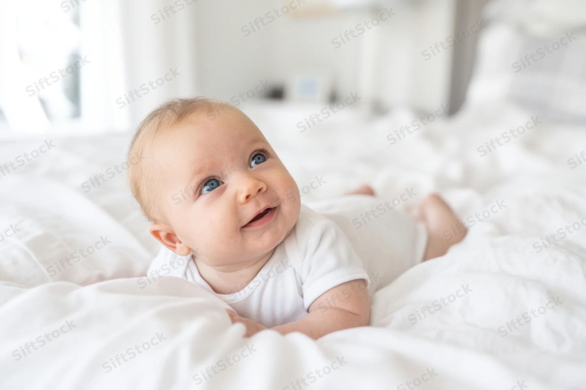 Baby lying on white bed in onesie gazing upward smiling