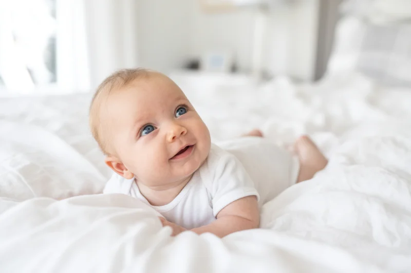 Baby lying on white bed in onesie gazing upward smiling