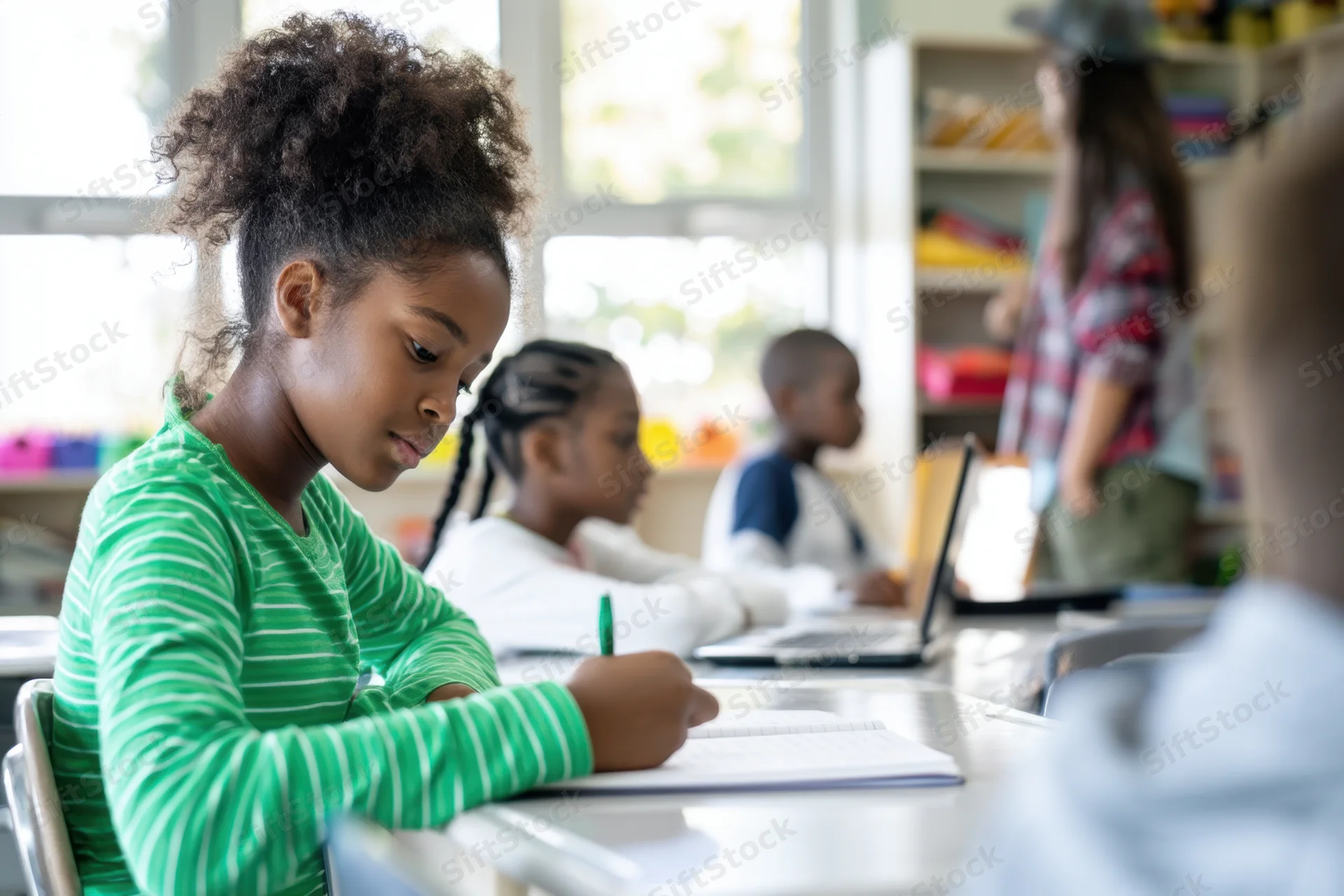 Young african girl writing in notebook at desk in bright classroom with classmates and teacher in background