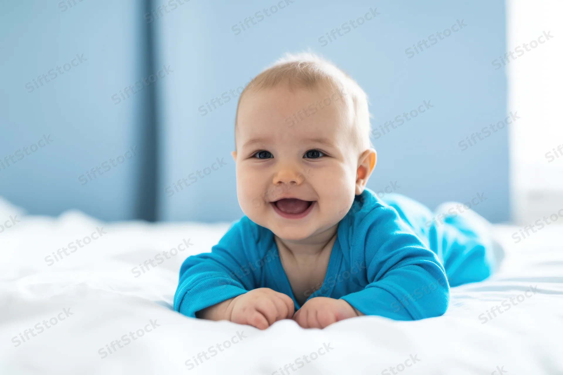 Baby smiling while lying on stomach on white bed wearing blue onesie