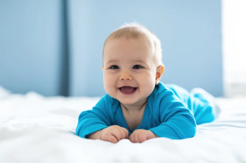 Baby smiling while lying on stomach on white bed wearing blue onesie