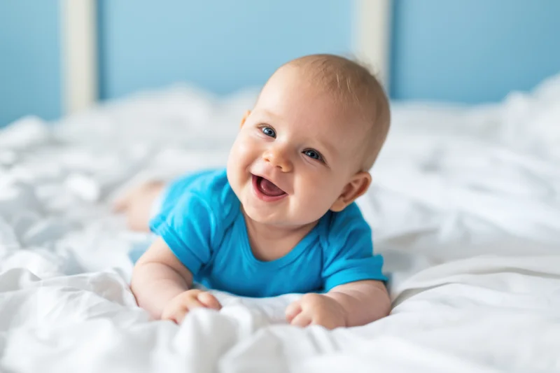 Baby smiling while lying on a white bed wearing a blue tshirt