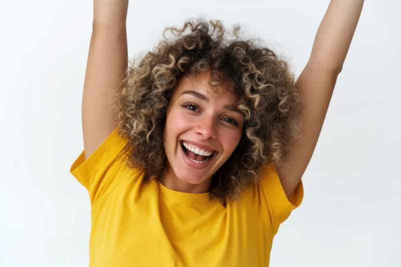 Cheerful woman with curly hair raising arms in joy, wearing yellow t-shirt against white background