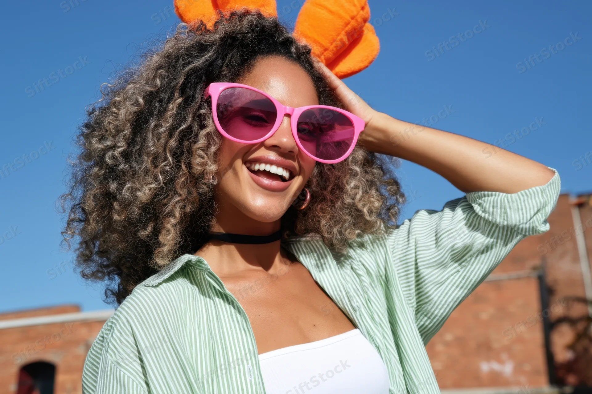 Cheerful young woman smiling with curly hair and pink sunglasses outdoors wearing orange bunny ears and striped shirt