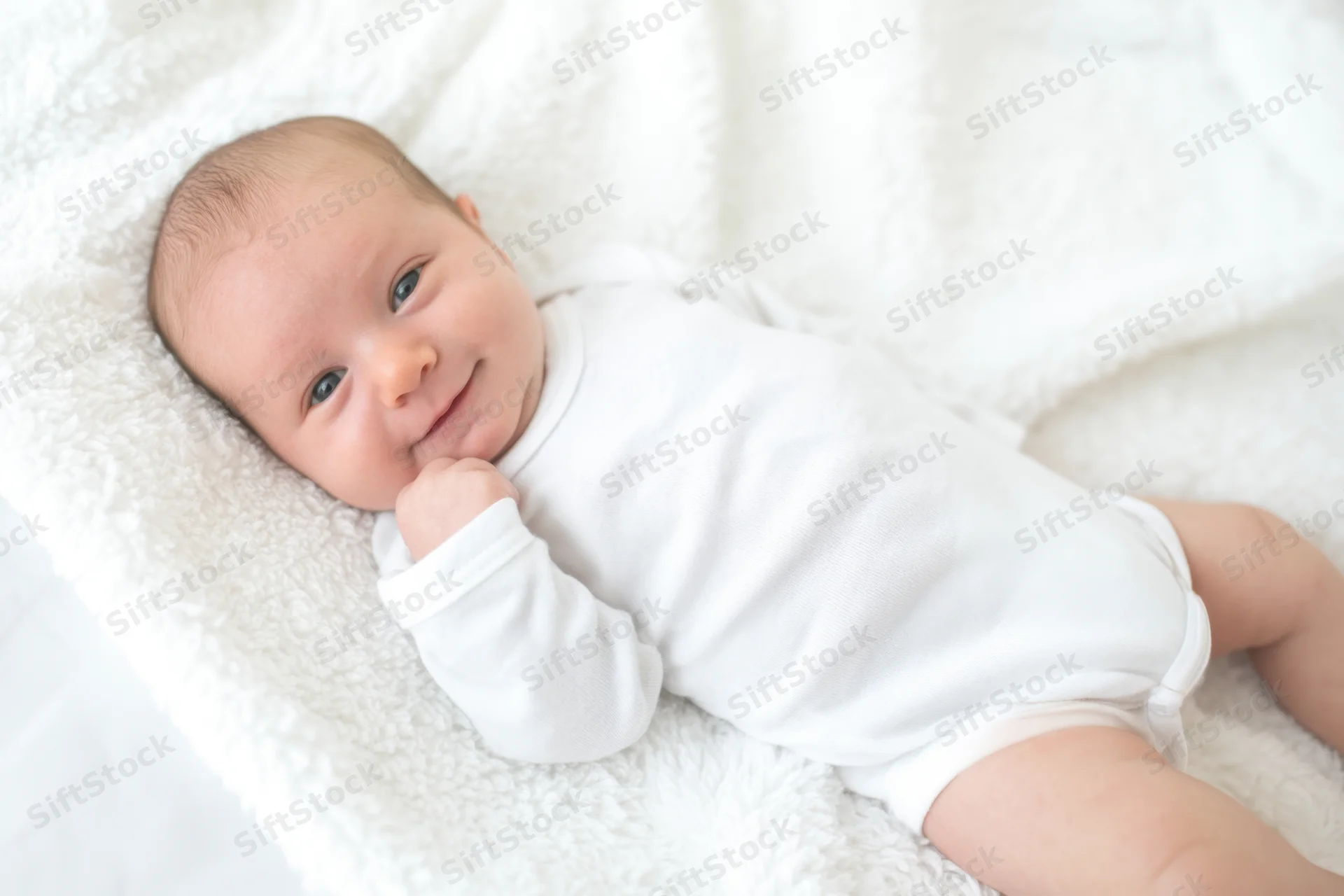 Newborn baby lying on a white plush blanket, smiling with hand near face
