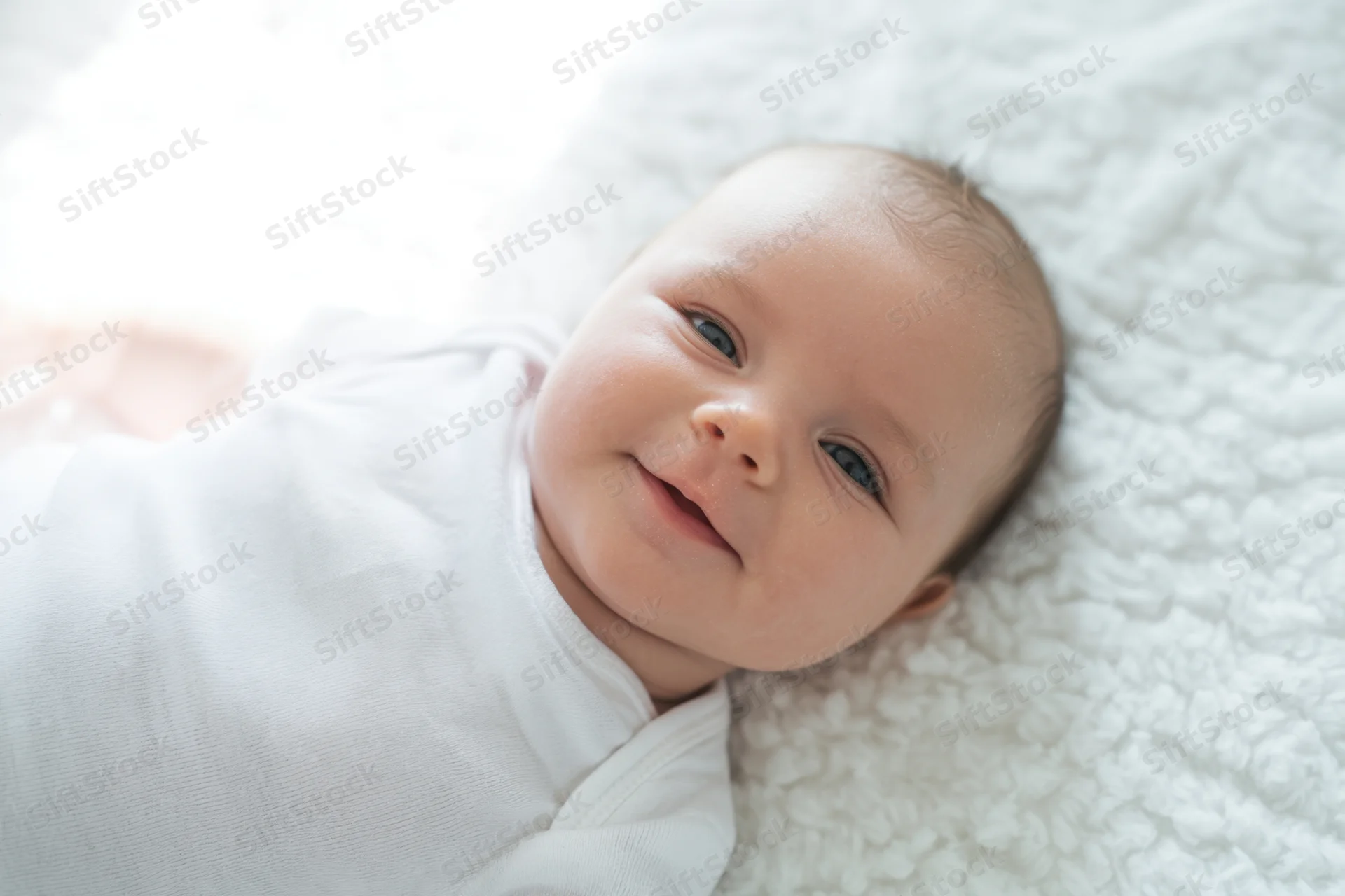 Smiling newborn baby lying on a soft white plush blanket in a bright indoor setting