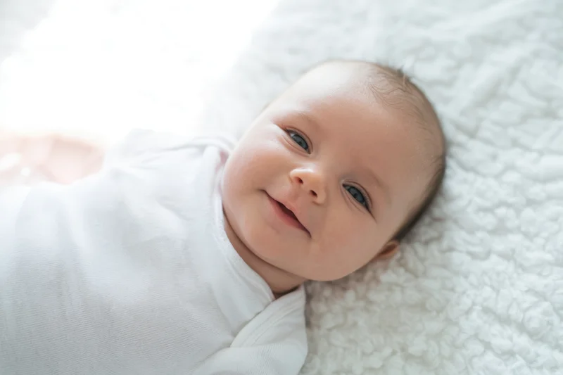 Smiling newborn baby lying on a soft white plush blanket in a bright indoor setting