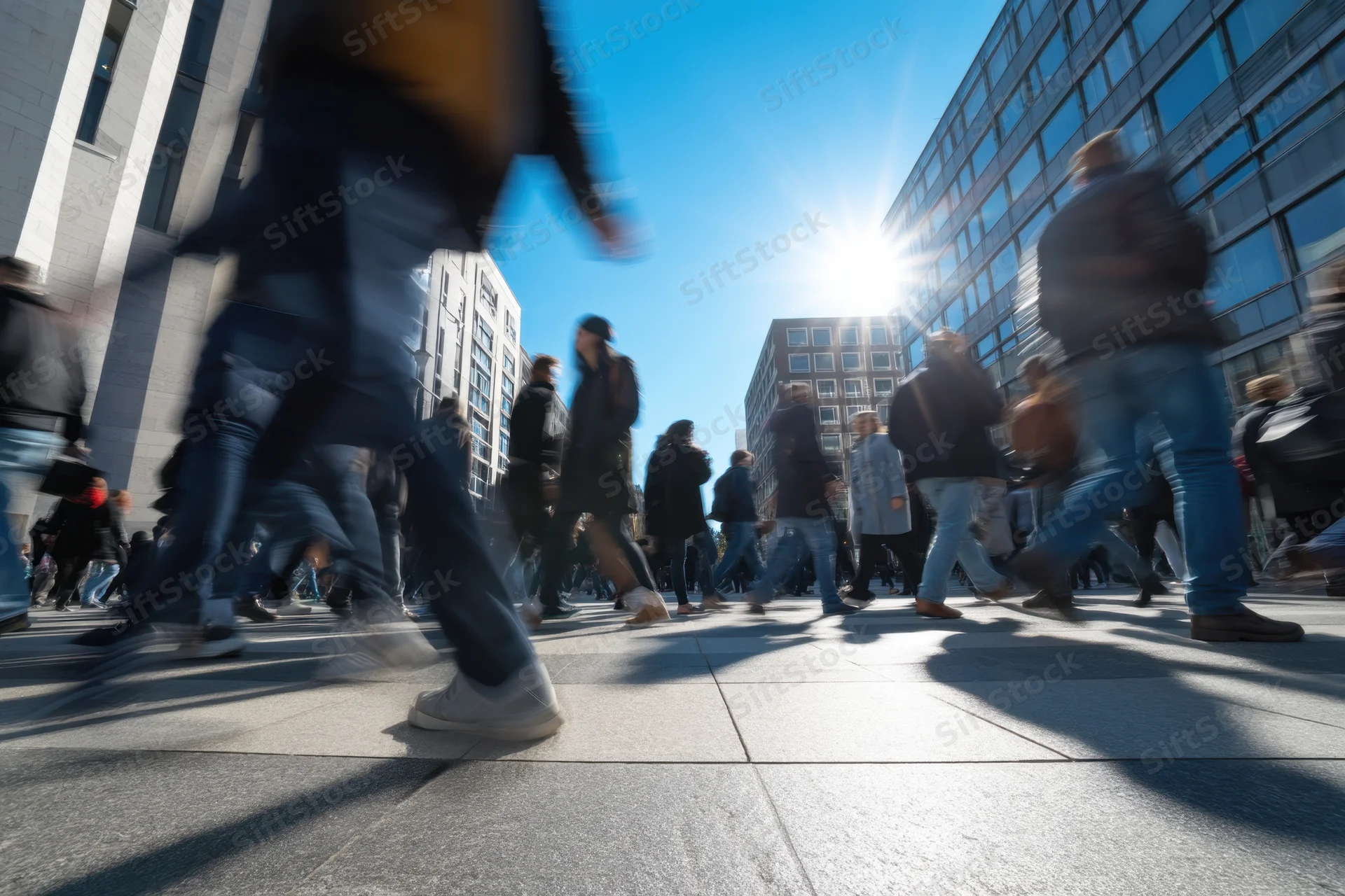 Blurred pedestrians crossing a city street in bright sunlight with modern buildings surrounding them